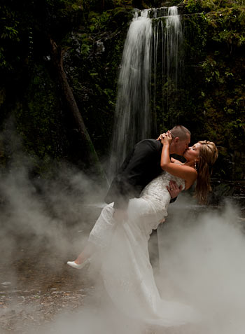 A rainforest wedding showing a bridal couple with a waterfall in the background, by wedding photographers melbourne. Waterfall Wedding Photography-the team from Wedding Photographers Melbourne feature a bridal couple enjoying a waterfall in a rainforest setting.
